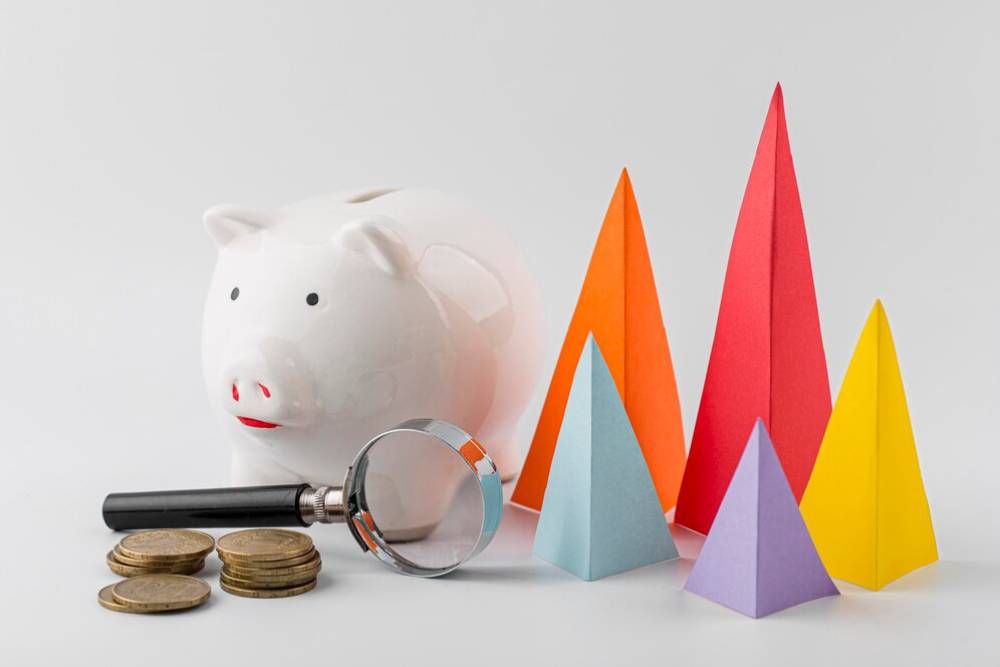 A white piggy bank beside a magnifying glass and stacked coins, with colorful paper pyramids in the background.