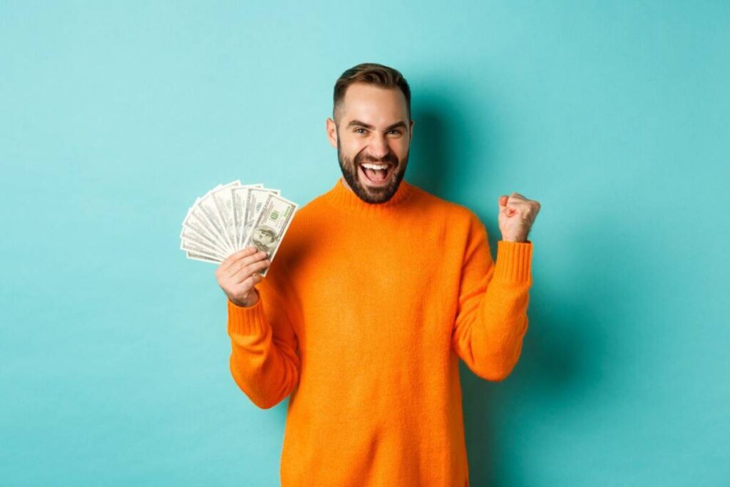 A person in an orange sweater holds a stack of cash and cheers, set against a bright blue background.