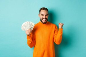 A person in an orange sweater holds a stack of cash and cheers, set against a bright blue background.