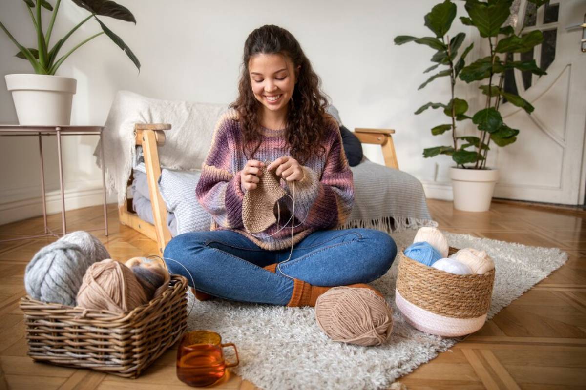 A woman sits on a cozy rug surrounded by skeins of yarn, knitting in a bright, plant-filled living space.
