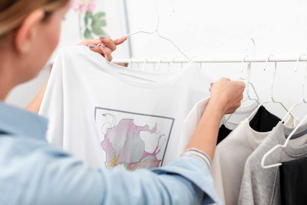 A person holds a white t-shirt with a colorful graphic design while browsing a selection of hanging clothes in a bright, stylish space.