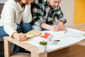 A cozy workspace featuring a miniature house model, sketch papers, and two individuals planning on paper.