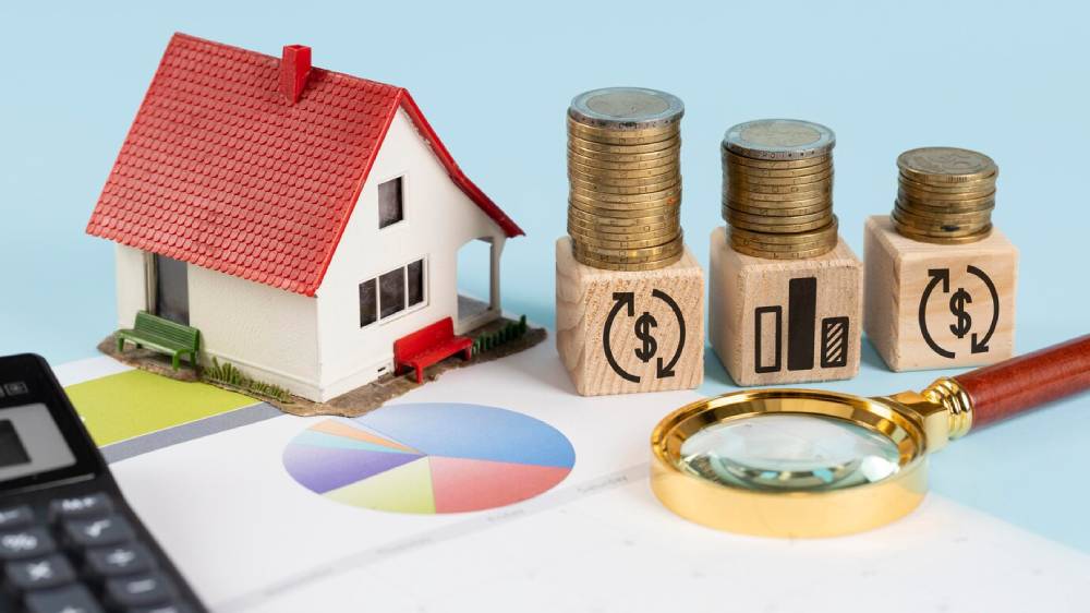 Model house beside stacked coins and wooden blocks with financial symbols, alongside a calculator and colorful pie chart.
