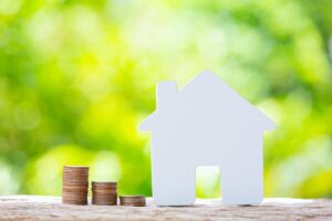 A white house figurine stands next to stacked coins, symbolizing real estate investment against a blurred green background.
