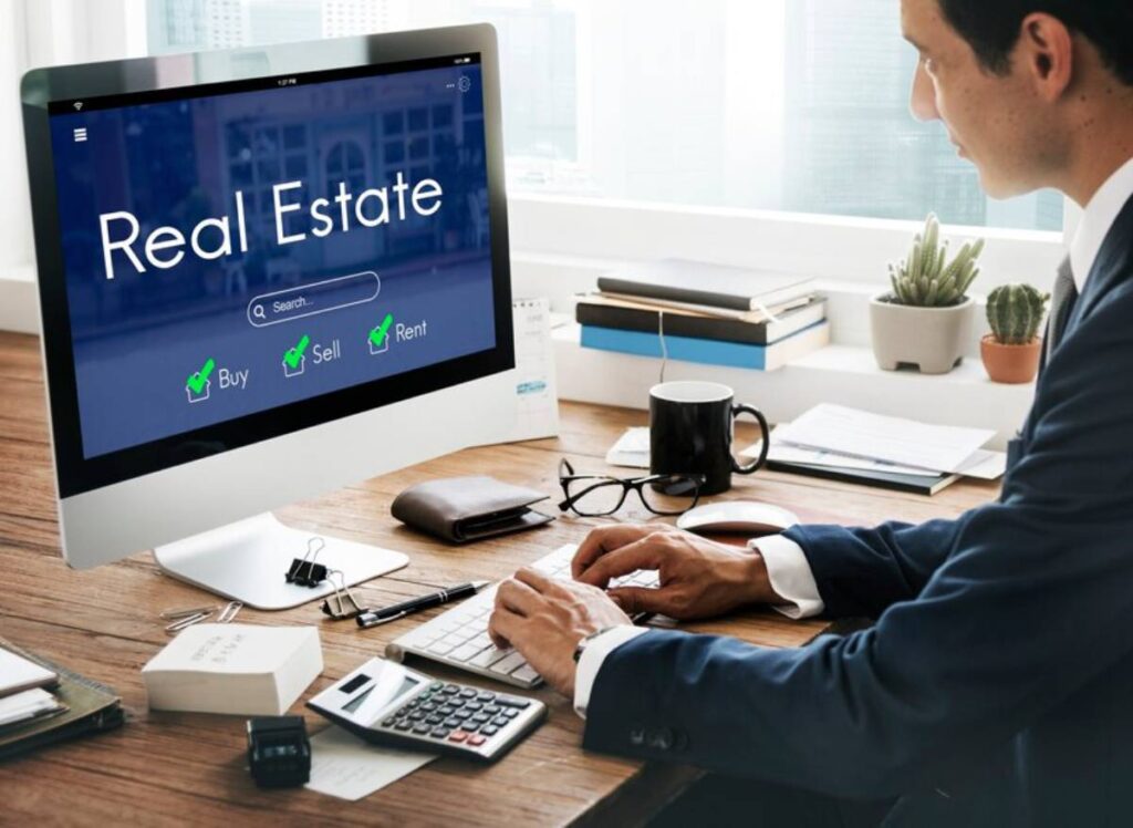A person in a suit using a computer displaying a real estate website with options to buy, sell, or rent, on a desk with office supplies.