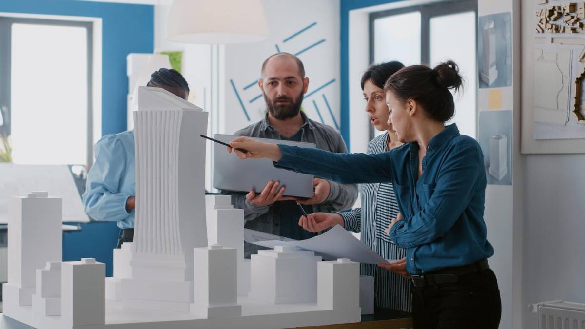 A woman gestures towards a large architectural model, while colleagues hold documents and discuss in a well-lit design studio.