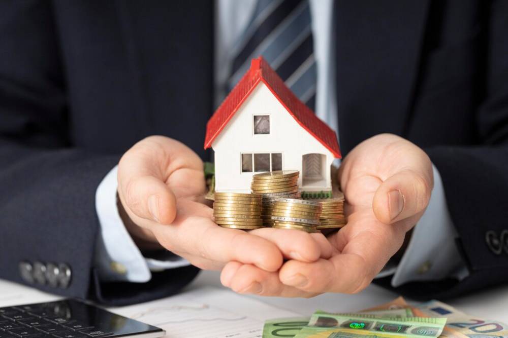 A businessman holds a small house model surrounded by stacks of coins, symbolizing real estate investment and financial growth./