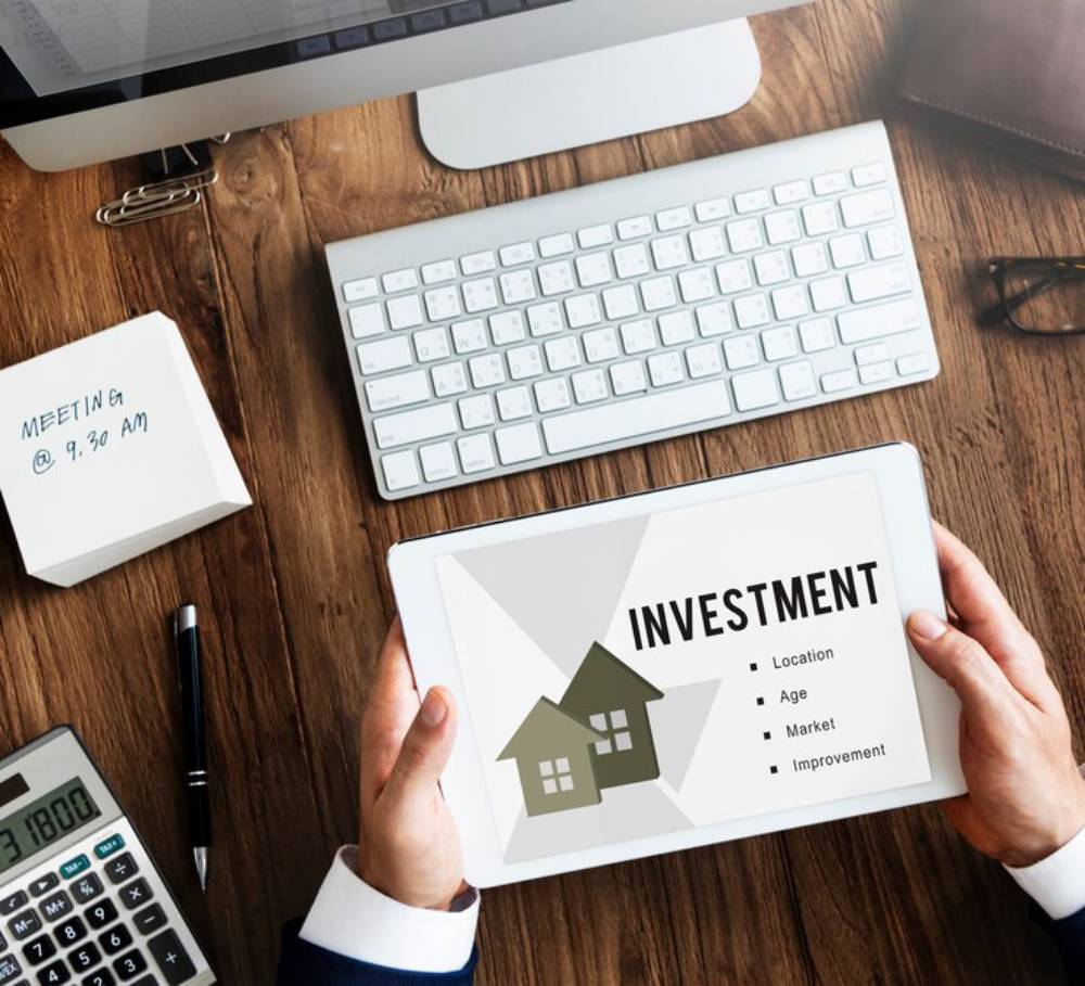 A person holding a tablet displaying a home investment strategy, with notes on location, age, market, and improvement on a desk.