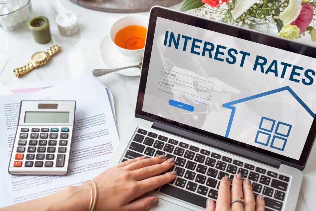 A person typing on a laptop displaying "Interest Rates" login page, with a calculator, documents on a table.