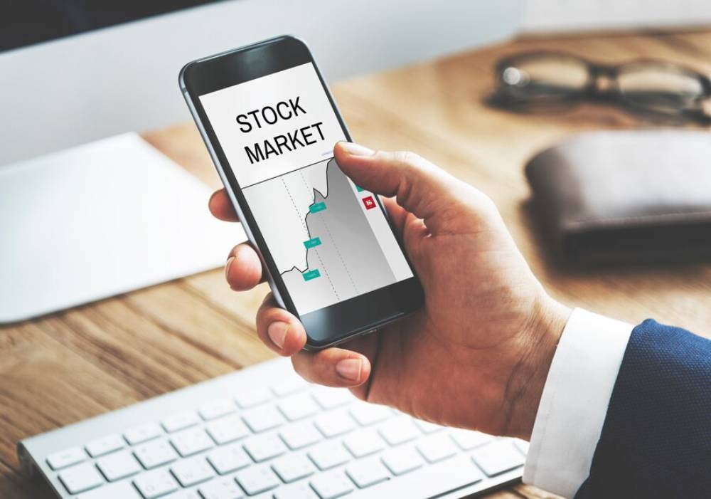 A person in a suit holds a smartphone displaying a stock market graph, with a keyboard and glasses in the background.