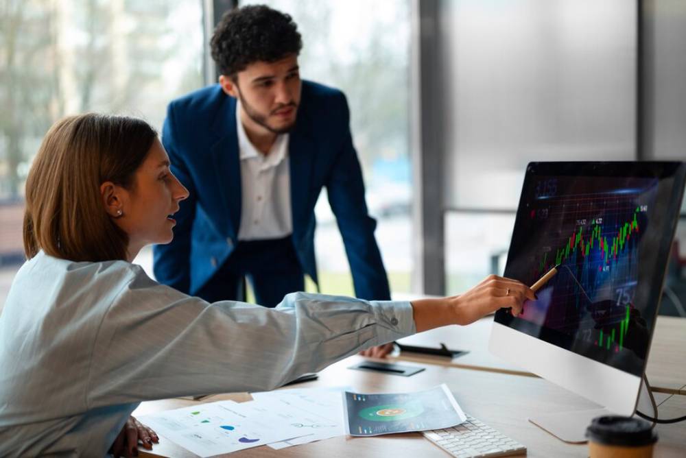 A woman points at a financial graph on a computer screen, while a man in a blue suit observes from behind in a modern office setting.