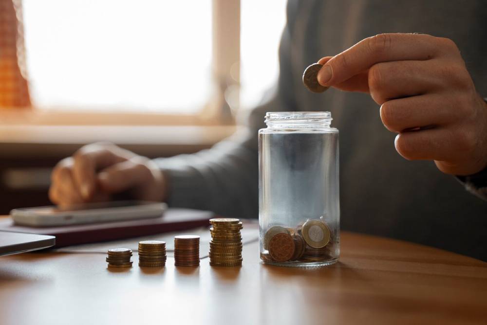 A person adding a coin to a clear jar filled with coins, with a stack of coins and a smartphone on a wooden table in soft light.