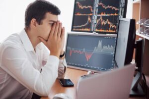 A man in a white shirt sits at a desk, stressed, with two monitors displaying risk and fluctuating cryptocurrency activity.