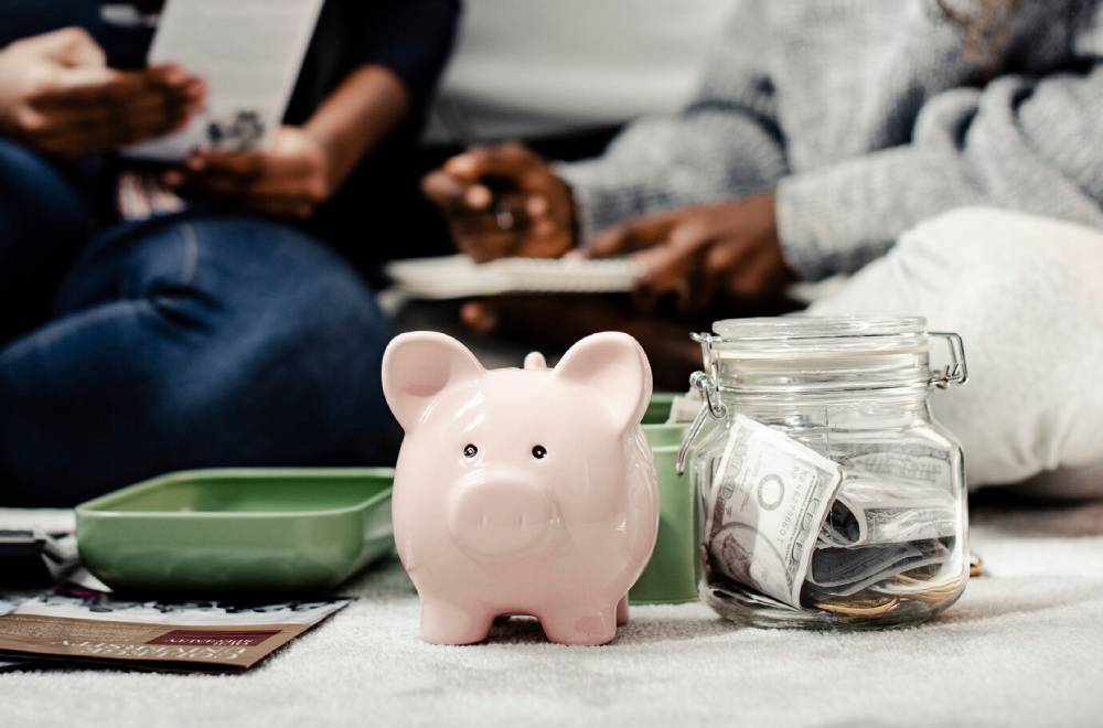 A pink piggy bank and a jar filled with cash sit on the floor, surrounded by people engaging in financial discussions.