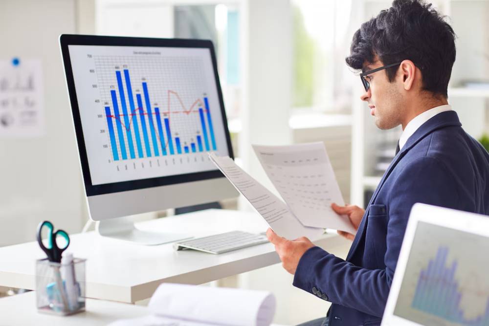 A man in a suit analyzes printed reports while looking at a computer screen displaying colorful graphs and data charts in an office.