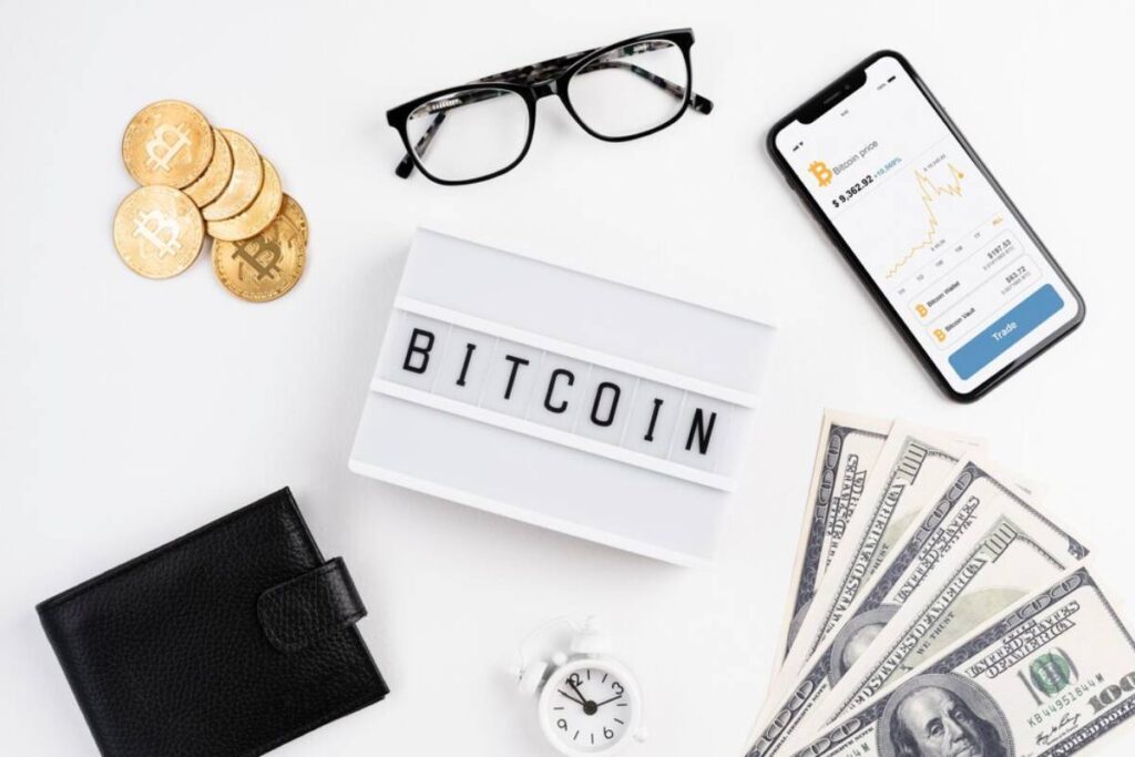 An arrangement featuring Bitcoin coins, glasses, a smartphone displaying market data, cash, a clock, and a "BITCOIN" sign.