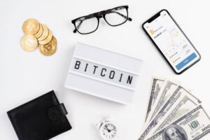 An arrangement featuring Bitcoin coins, glasses, a smartphone displaying market data, cash, a clock, and a "BITCOIN" sign.