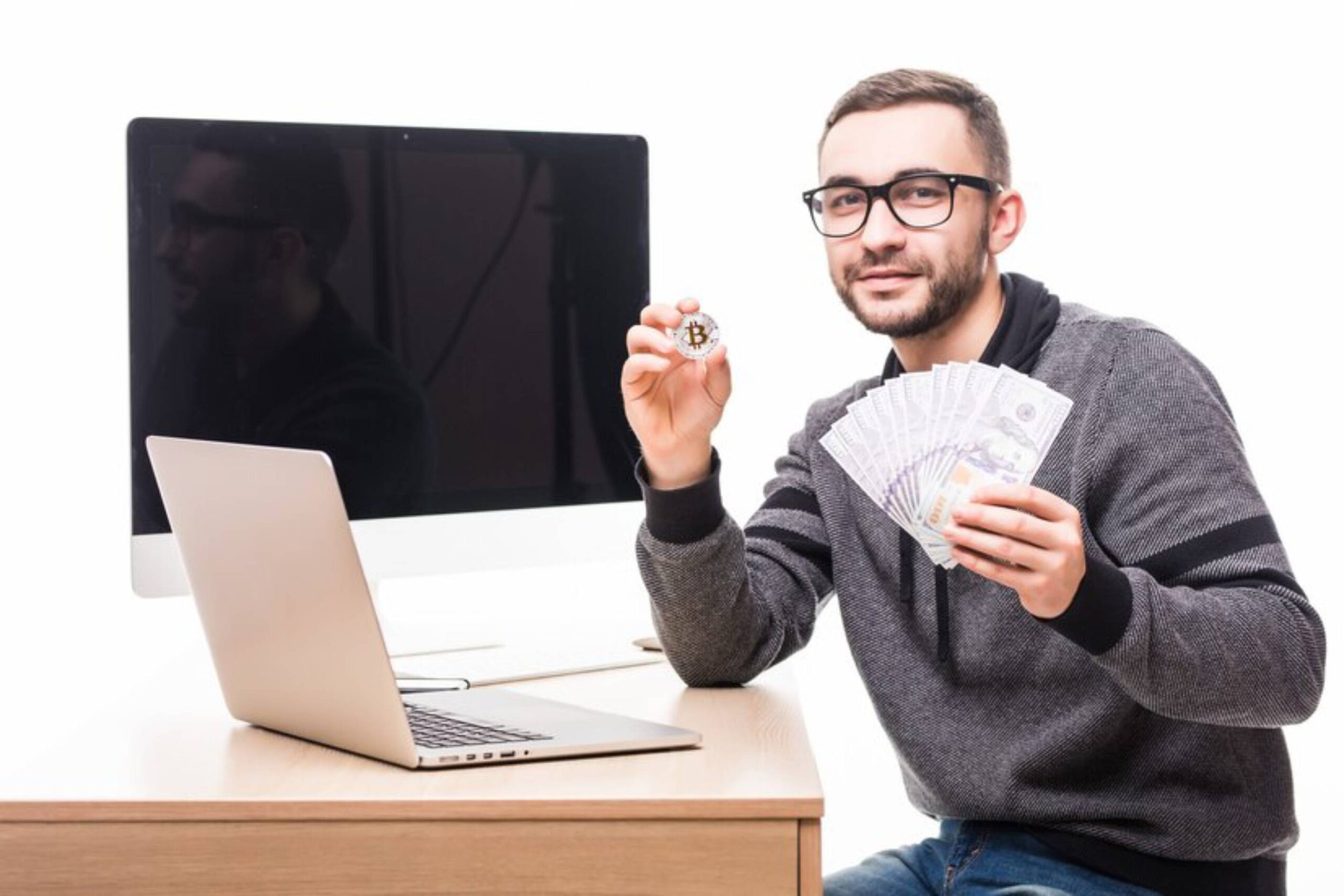 A person holding a Bitcoin and bundles of banknotes, sitting at a desk with a laptop and a large monitor in the background.