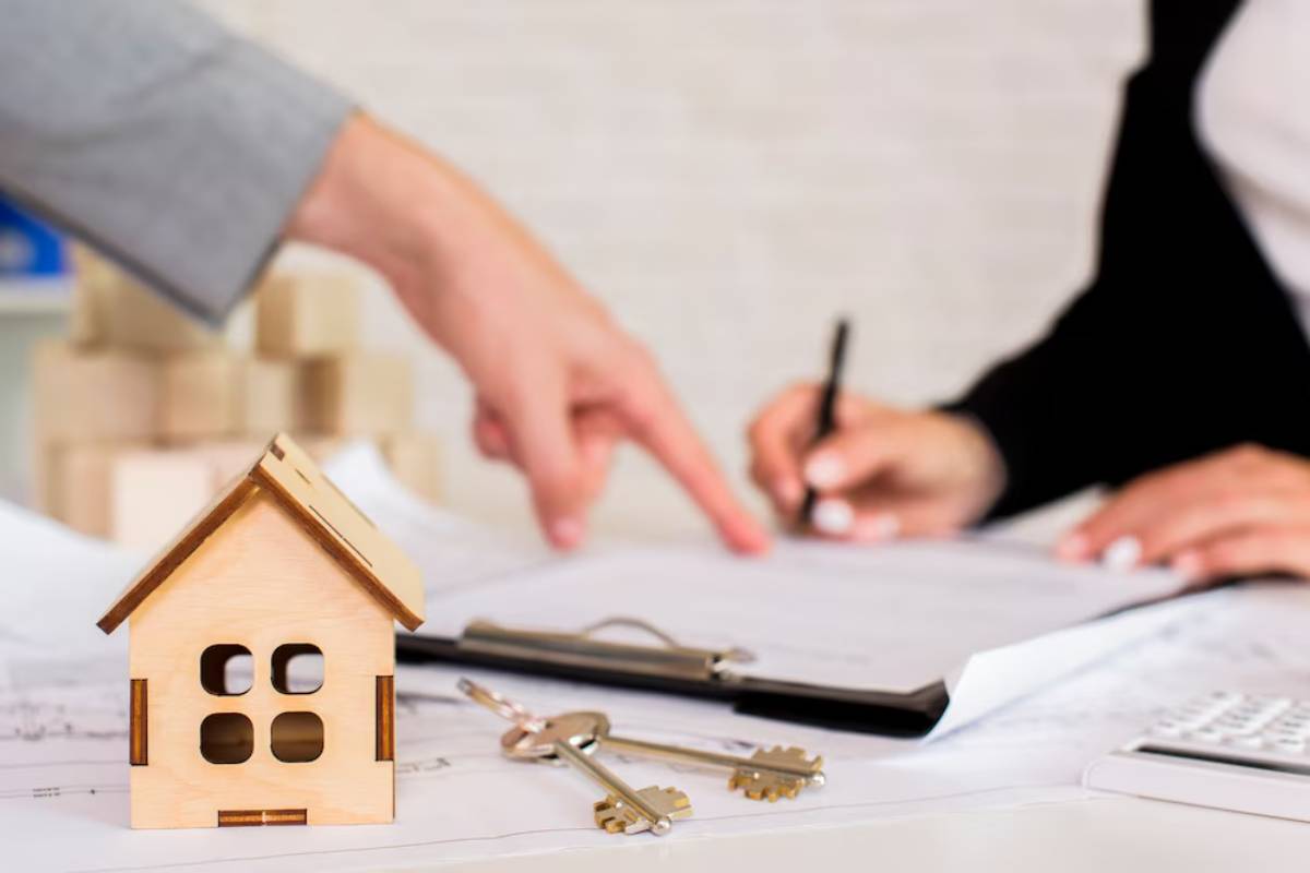 A small wooden house model with keys on plans as two people discuss paperwork, indicating a real estate transaction.