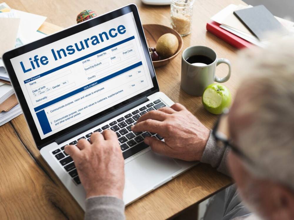 An elderly man fills out a life insurance form on his laptop, surrounded by fruits and a coffee cup on a wooden table.