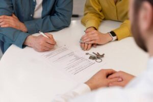 A couple is seated at a table, reviewing and signing a document while keys rest beside them, indicating a property transaction.