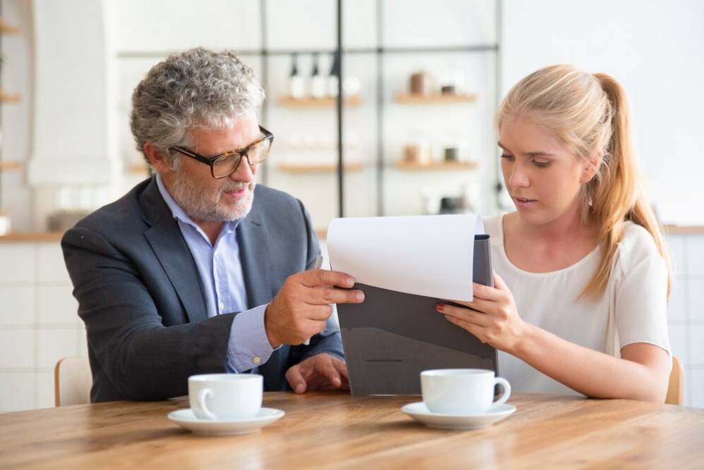 A man in a suit discusses a document with a woman in a white top at a wooden table, two coffee cups in front of them.